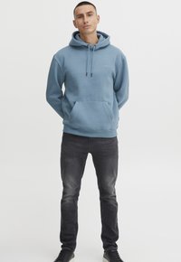 Felpa azzurra chiarissima con cappuccio e lacci, accompagnata da jeans grigi scuri e sneakers nere, mostrata su uno sfondo semplice.
