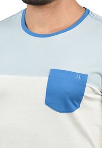 Bomull T-skjorte med lys blå og hvit fargeblokkdesign, rund hals, og en kontrasterende blå brystlomme med detaljert søm.