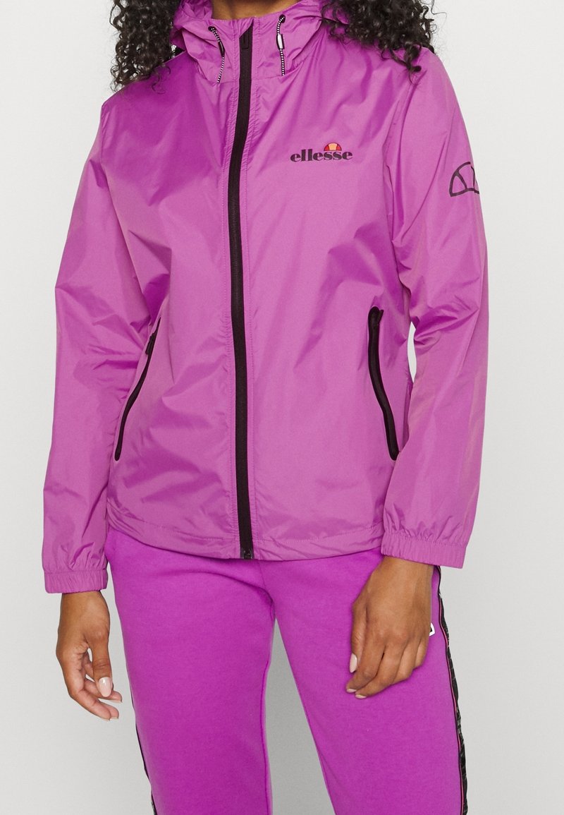 Kvinna i en lila ellesse jacka med huva, svart dragkedja och fickdetaljer, matchad med magenta byxor med svarta sidostråkar.