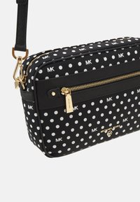 Czarna torebka crossbody w białe kropki, ze złotymi akcentami, płaski design oraz logo. Kieszeń zapinana na zamek z przodu.