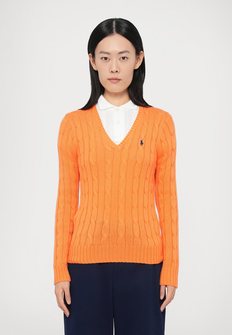 Oranžs džemperis ar adījumu rakstu, V-veida izgriezumu, ribotiem manžetiem un apakšu. Uz krūtīm ir neliels logotips. Valkāts virs baltas kreklam līdzīgas krekla.