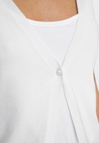 Cardigan bianco lavorato a maglia con scollo a V, caratterizzato da una chiusura a singolo bottone di perla e bordi a coste. Indossato sopra una canottiera bianca.