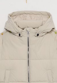 Chaqueta acolchada beige con capucha, cierre frontal, botones a presión negros y dos bolsillos laterales. Incluye un texturizado acolchado y acentos de botones coloridos.