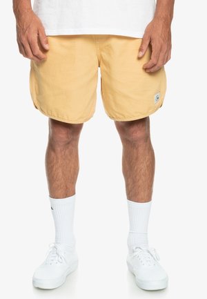 Homme portant un short jaune pâle, des chaussettes blanches montantes et des baskets blanches, debout les mains détendues le long du corps.