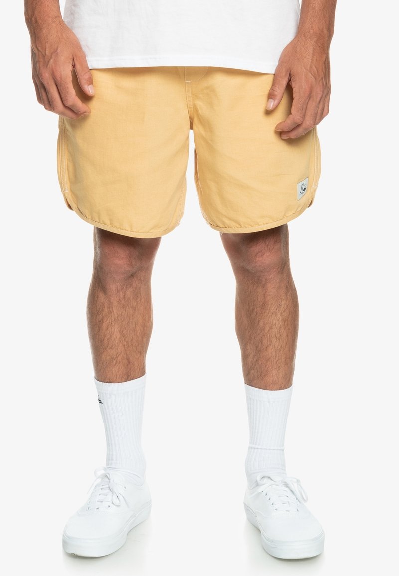Homme portant un short jaune pâle, des chaussettes blanches montantes et des baskets blanches, debout les mains détendues le long du corps.