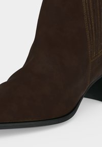 Gros plan d'une botte en cuir marron avec des coutures visibles et une semelle et un talon noirs sur fond blanc.