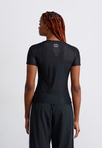 Woman with long red braids wearing a fitted black short-sleeve shirt and loose black pants with side buttons, facing away against a plain background.