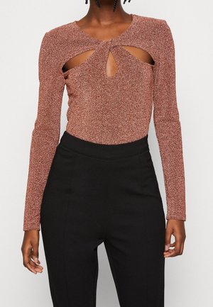 Henkilö, joka on pukeutunut pitkähiharaiseen ruosteenväriseen kimaltelevan topin, jossa on tipan muotoisia aukkoja rinnassa, yhdistettynä korkeavyötäröisiin mustiin housuihin.