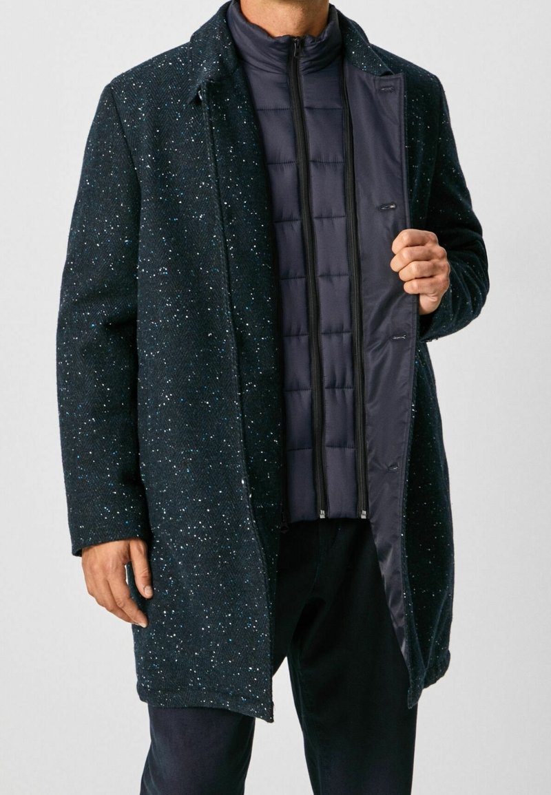 Kappa i ullblandning i marinblått med en fläckig textur och uppvikt krage, lagd över en mörk vadderad innerjacka med dragkedja.