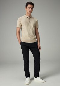 Beige-paitainen polo, lyhythihainen ja kolmella mustalla napilla, yhdistetty mustiin housuihin ja valkoisiin lenkkitossuihin. Suora rakenne, rento istuvuus.