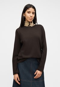 Νεαρή γυναίκα με μακριά σκούρα μαλλιά φοράει μαύρο μακρυμάνικο τοπ πάνω από μπεζ ζιβάγκο με σχέδια και μπλε τζιν φούστα, στέκεται μπροστά σε λευκό φόντο.