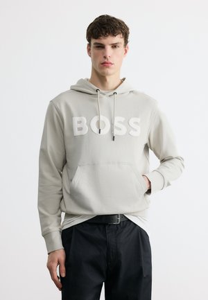 Jeune homme portant un sweat à capuche gris clair avec le texte « BOSS », ceinture noire et pantalon noir, debout devant un fond blanc uni.