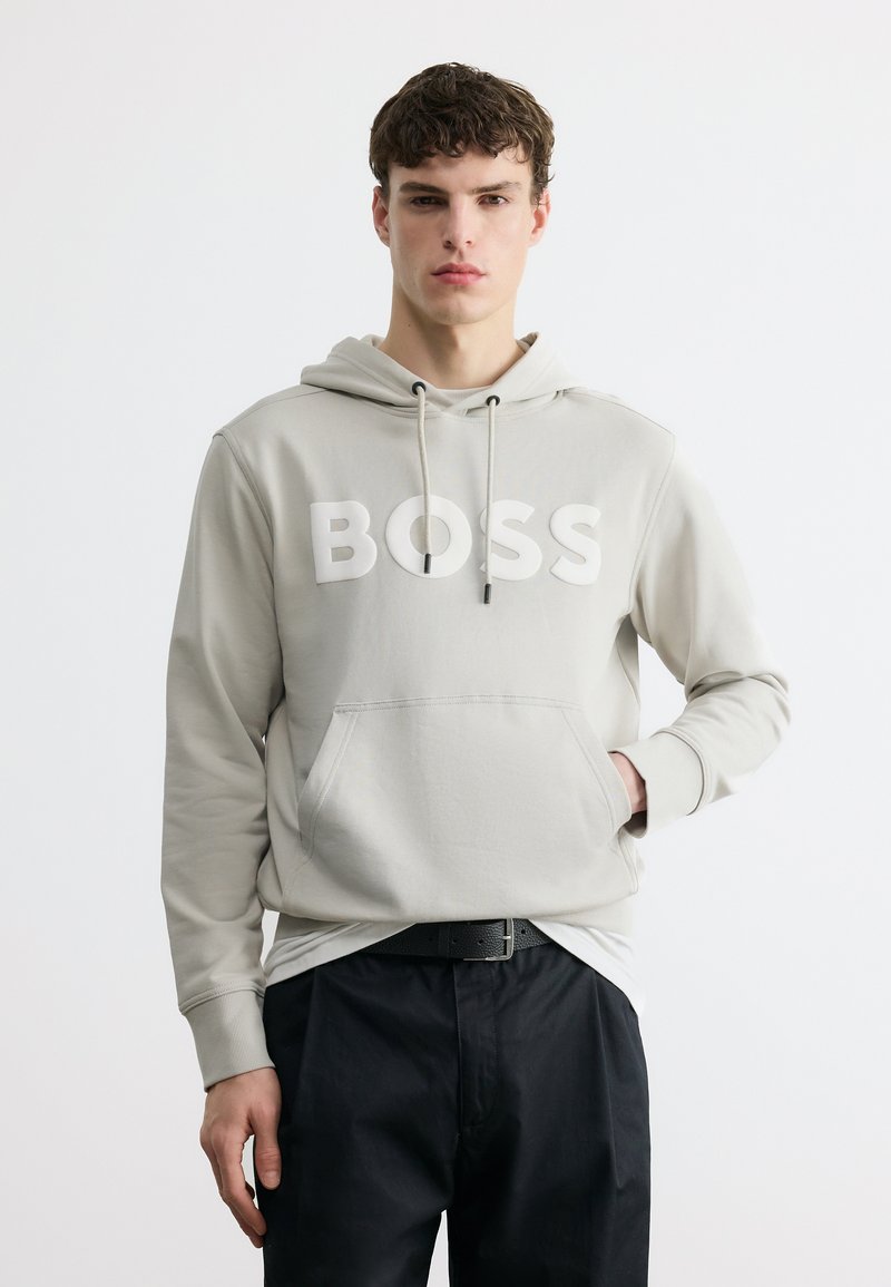 Jeune homme portant un sweat à capuche gris clair avec le texte « BOSS », ceinture noire et pantalon noir, debout devant un fond blanc uni.