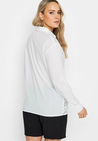 Blusa blanca de manga larga con detalles de ojales en los hombros y el dobladillo, corte holgado y cierre de botones en los puños.
