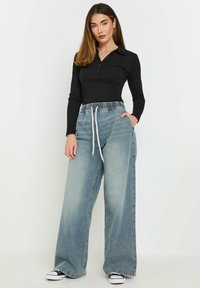 Top lungo nero a maniche lunghe con colletto bottonato, abbinato a jeans ampi a vita alta di colore blu chiaro, con cintura elastica e lacci.