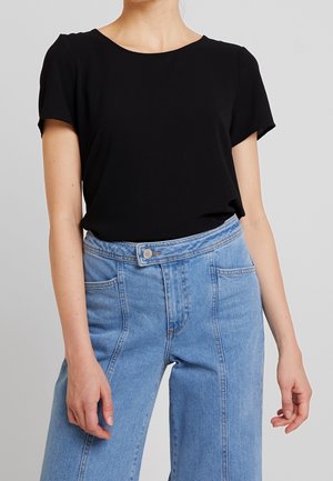 Femme portant un haut noir uni à manches courtes, rentrée dans un jean en denim bleu clair taille haute avec des détails de couture à l'avant.