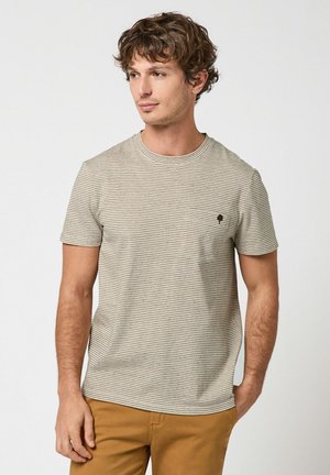 Homme aux cheveux bouclés portant un t-shirt beige clair à fines rayures et un pantalon couleur moutarde, regardant vers la droite avec une main dans la poche.
