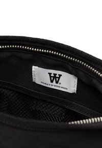 Intérieur d'un sac en tissu noir avec une fermeture éclair argentée, comportant une étiquette blanche avec la marque "DOUBLE V BY WOOD WOOD" et une poche en maille texturée.