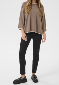 Femme portant un pull ample taupe, un pantalon skinny noir et des mocassins à plateforme noirs sur un fond blanc uni.