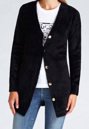Femme portant un cardigan noir en peluche boutonné sur un t-shirt blanc à imprimé graphique et un jean bleu, debout, les mains détendues le long du corps.