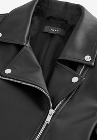 Zwarte neplederen jas met een scherpe revers, zilveren rits en drukknoopdetails, met een gladde textuur en een op maat gemaakte pasvorm. Merkenlabel zichtbaar.