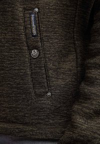 Primo piano di una tasca di giacca di colore marrone scuro con una texture, adornata da due bottoni a pressione in metallo e un'etichetta in tessuto verticale che riporta la scritta "Schmuddelwedda".