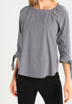 Vrouw in een zwart-wit gestreepte blouse met lange mouwen en strikmanchetten, en een zwarte broek, staand tegen een effen achtergrond.