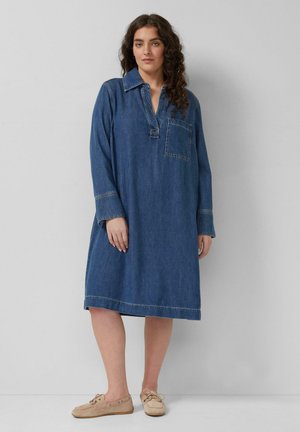 Kobieta z długimi kręconymi włosami, ubrana w niebieską sukienkę dżinsową do kolan z długimi rękawami oraz beżowe buty casual, stoi na tle szarej ściany.