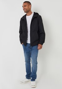 Sort hættejakke med to lynlåslommer på brystet, båret over en hvid T-shirt, parret med blå jeans og hvide sneakers.