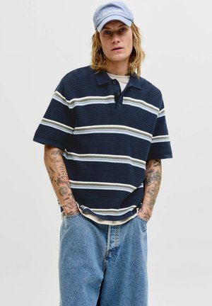 Junge Person mit langen Haaren, die ein blau-weiß gestreiftes Polohemd, eine hellblaue Kappe, lockere blaue Jeans und sichtbare Arm-Tattoos trägt.