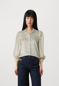 Krémová blúzka s čiernym abstraktným bodkovaným vzorom, s predným zapínaním na gombíky a volánovými rukávmi. Nosená s tmavými džínsami. Hladká, ľahká tkanina.