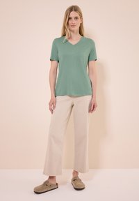 T-shirt verde chiaro a scollo a V realizzato in tessuto morbido, abbinato a pantaloni beige a gamba larga. Il modello indossa zoccoli in suede marrone. Outfit semplice e casual.