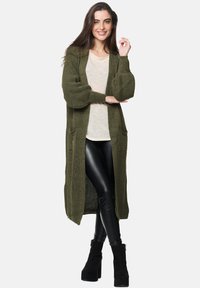 Langer olivgrüner Cardigan mit strukturiertem Strick, offener Front und großen Taschen. Kombiniert mit einem hellen Oberteil und schwarzen Lederhosen.