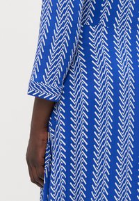 Blau gemustertes Kleid mit weißem Zickzackdesign, ausgestattet mit einem aufgerollten Ärmel und glatter Stofftextur. Sichtbare Hand an der Seite.