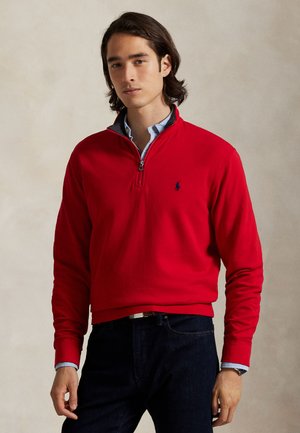 Jeune homme aux cheveux mi-longs foncés portant un pull rouge à fermeture éclair quart de zip, une chemise bleu clair en dessous, un jean foncé et une montre au poignet gauche.