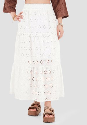 Femme portant une jupe midi blanche ajourée, des sandales marron à motifs, et un haut marron à manches longues avec des bracelets et une montre.