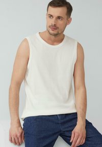 Weißes ärmelloses Shirt aus weichem Stoff, mit rundem Halsausschnitt und glatter Textur, kombiniert mit dunkelblauen Jeans.