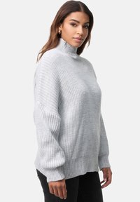 Jasnoszary sweter z dzianiny z wysokim kołnierzem, prążkowanymi mankietami i dołem; luźny krój z teksturowanym wzorem. Model nosi czarne dżinsy.