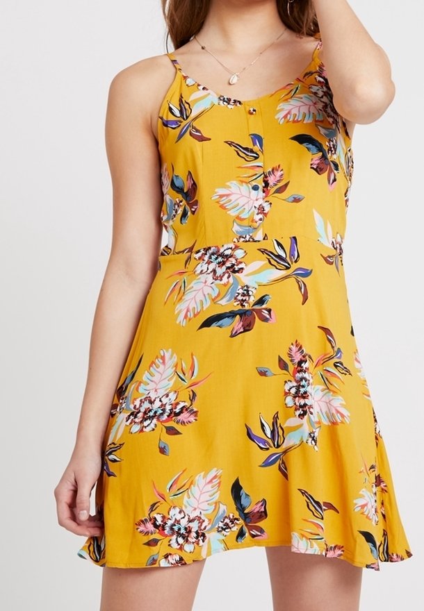 Femme portant une robe jaune sans manches avec des motifs floraux roses, bleus et blancs, ainsi qu'un collier délicat avec un petit pendentif.