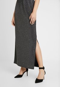 Robe maxi noire pailletée avec une fente sur le côté, présentant un corsage ajusté et un tissu doux et texturé. Associée à des chaussures à talons hauts noires et à bout pointu.