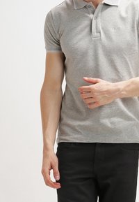 Šedé polo tričko s krátkými rukávy, límečkem a zapínáním na dva knoflíky. Obsahuje kontrastní lemování kolem límce a rukávů. Nosené s černými kalhotami.