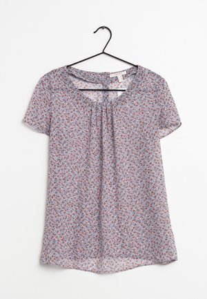 Blusa leggera azzurro chiaro a maniche corte con piccolo motivo floreale rosso e bianco, scollatura tonda con parte anteriore plissettata, appesa a un appendiabiti nero.