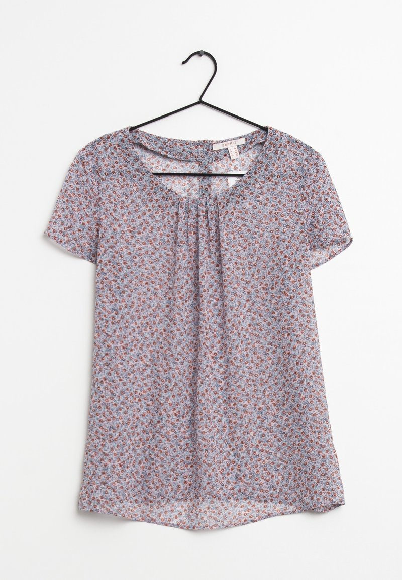Blusa leggera azzurro chiaro a maniche corte con piccolo motivo floreale rosso e bianco, scollatura tonda con parte anteriore plissettata, appesa a un appendiabiti nero.