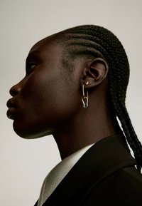 Pendiente de aro dorado con un acento de estrella, llevado en la oreja. El sujeto tiene el pelo trenzado y viste una chaqueta negra con un collar de satén.