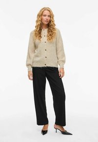 Beige stickad kofta med puffärm, knappar fram och borstad yta, kombinerad med svarta vida byxor och svarta spetsiga klackar.