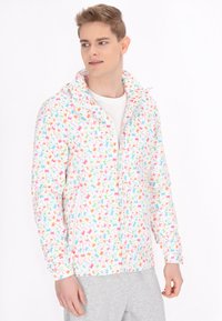 Giacca leggera con zip su sfondo bianco e motivo a foglie colorate in rosa, blu e verde. Presenta un colletto alto e tasche laterali.