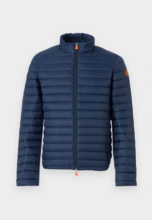 Marinblå pufferjacka med horisontell sömnad, hög krage och en framzip som har en orange dragflik. Prydd med en logotyp på ärmen.