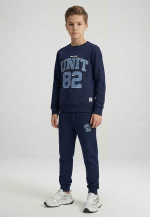 Jongen staand, gekleed in een marineblauwe trui en broek met "UNIT 82" print, en witte sneakers, op een effen lichte achtergrond.