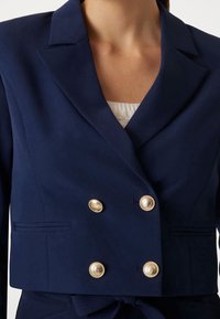 Marineblauwe dubbelgeknoopte blazer met gouden knopen, puntige revers en zijzakken; gemaakt van een soepele stof met een getailleerde pasvorm.