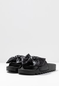 Black slide sandals with thick textured soles and large, padded bow straps across the top.